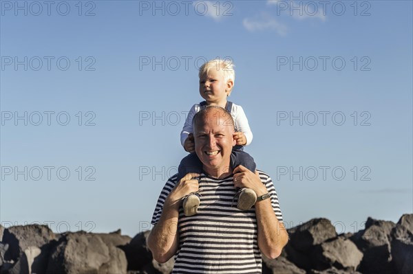 Happy father holding his small blond son on his shoulder against blue sky