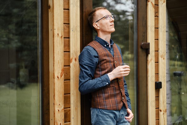 Pensive middle age man stands on the terrace of a cozy bungalow and drinks bourbon