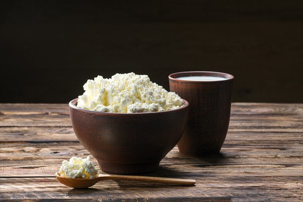 Fresh cottage cheese in clay bowl with wooden spoon with a glass of milk on rustic wooden background