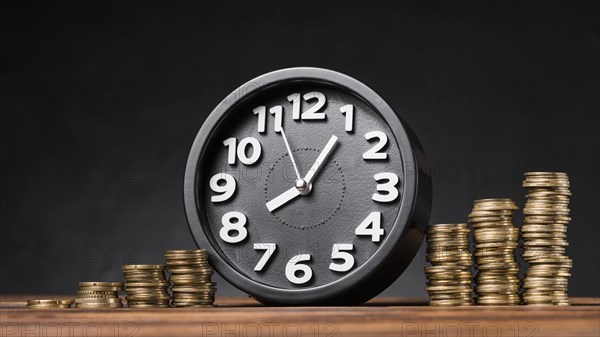 Round clock between the increasing coins on wooden desk against black backdrop
