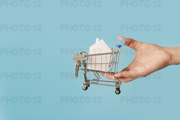 Close up hand holding miniature shopping cart with paper house keys against blue background