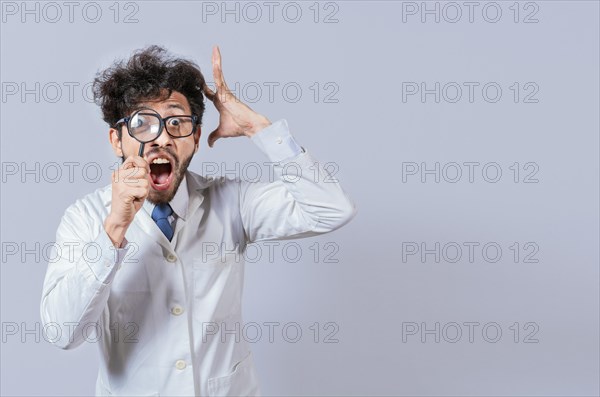 Surprised male scientist in lab coat looking at camera with magnifying glass. Mad scientist holding a magnifying glass isolated