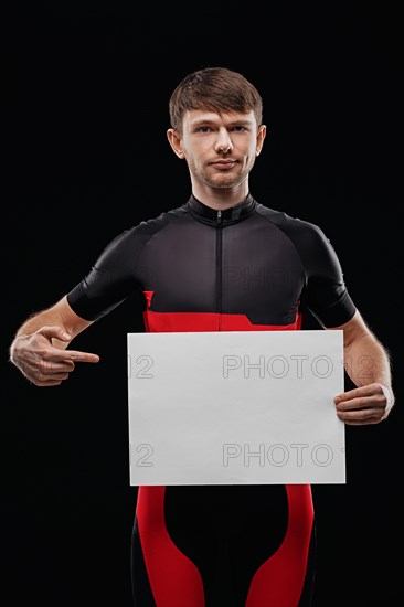 Sport. Cyclist in training clothes on black background holding blank sheet of paper. Your text here