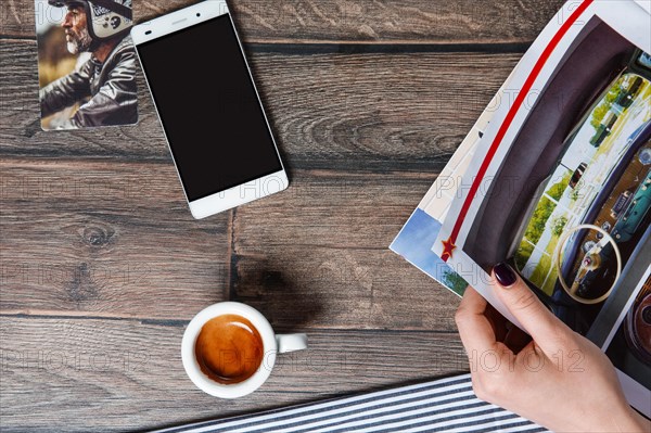 Top view of table with cup of coffee and smartphone. Hand holding magazine