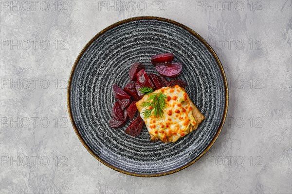 Top view of fried cod fillet with melted cheese topping and roasted beetroot slices on grsy stone background