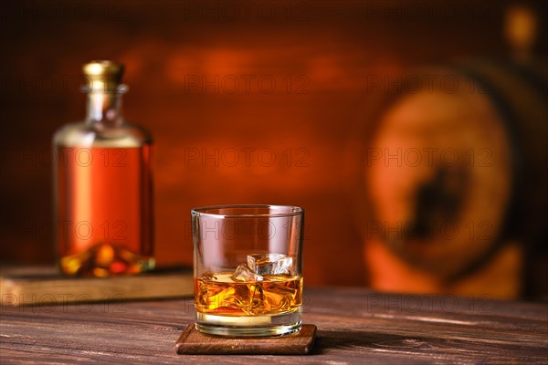 Glass of whiskey with ice on wooden table with bottle and barrel on background
