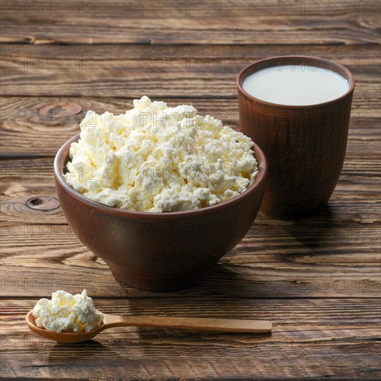 Fresh cottage cheese in clay bowl with wooden spoon with a glass of milk on rustic wooden background