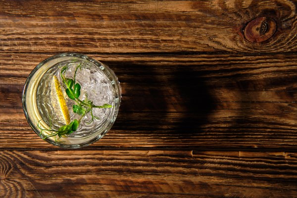 Overhead view of glass with cold water with ice and lemon casting a shadow on wooden table