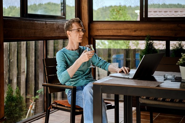 Middle aged man drinking wine and chatting in messenger on laptop