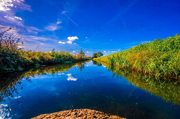 A small irrigation canal between two fields with reflection of clouds and the blue sky in autumn