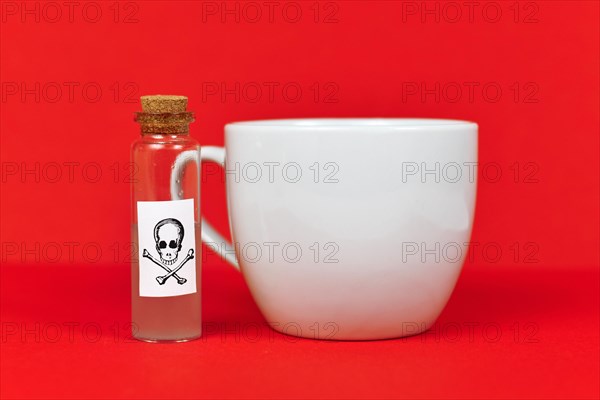 Vial with cork and poison skull label next to white tea cup on red background