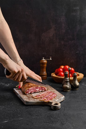 Female hand with knife slicing smoked pork sausage in organic casing on wooden cutting board