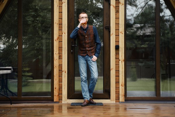 Pensive middle age man stands on the terrace of a wooden bungalow and drinks bourbon