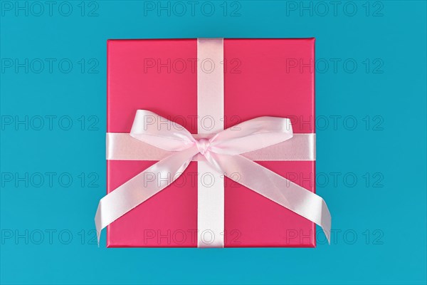 Single square shaped pink gift box with tied ribbon bow on teal blue background