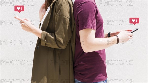 Couple standing back back using cellphone with love messages icons against wall