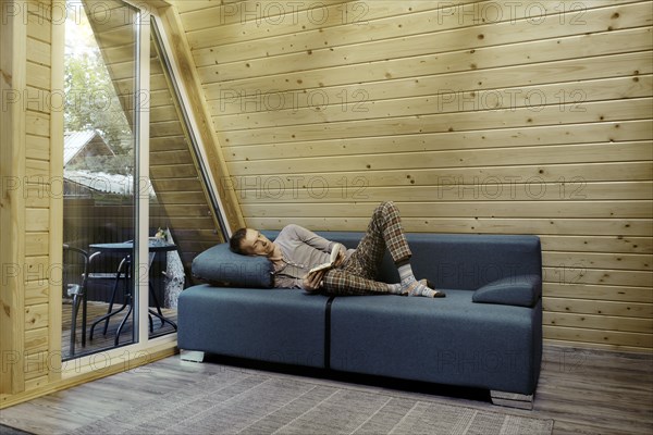 Middle aged man has retired to a country wooden cabin and is reading a book lying on the sofa