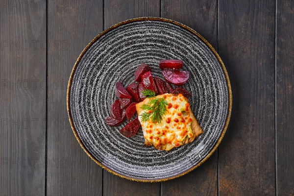 Top view of fried cod fillet with melted cheese topping and roasted beetroot slices on dark wooden table