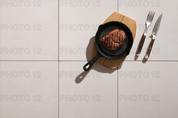 Top view of grilled beef steak in cast iron skillet on ceramic tile background with harsh shadow