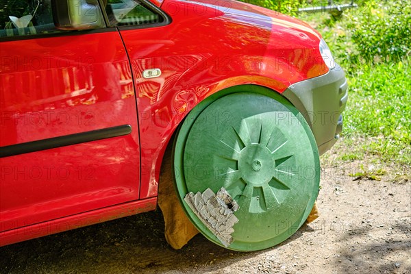 A red car with a green lid of a rain barrel at the place where the right front wheel should be