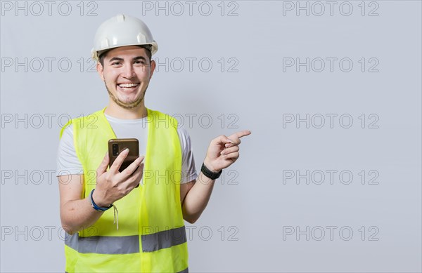 Happy Civil engineer holding cellphone and pointing a promo. Happy engineer holding phone and pointing to the side isolated. Young construction engineer holding phone and pointing at promo