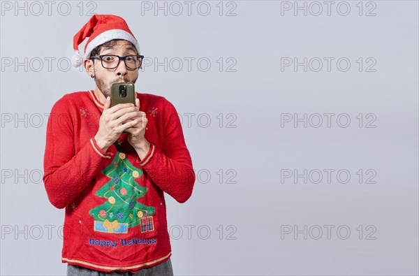 Man in christmas hat with amazed face holding phone. Online Christmas special offers concept. Young man in christmas clothes and hat amazed holding the phone