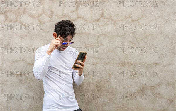 Surprised young Latin looking at his cell phone leaning against a wall. Handsome guy in glasses amazed with his cell phone leaning against a wall