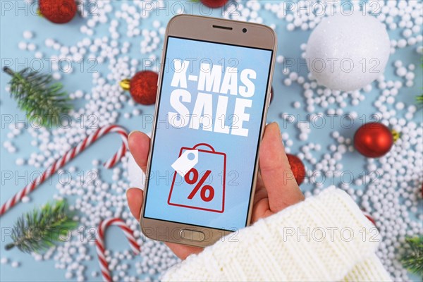 Concept for Christmas seasonal online shopping and sales wit hand holding cell phone with X-Mas Sale sign in front of desk with seasonal decorations