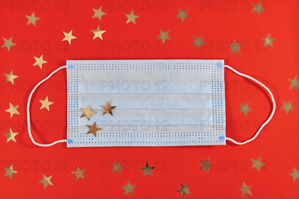 Christmas during Corona Virus times concept with medical face mask surrounded by golden star ornaments on red background