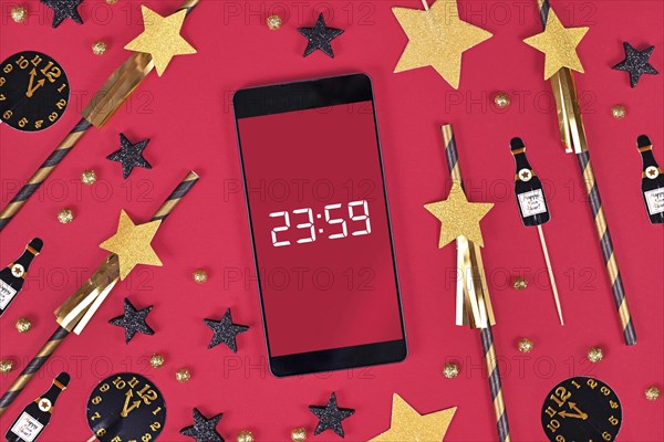 New Year Silvester celebration concept with smart phone with timer countdown to midnight between party items like paper straws and paper champaign bottles on dark pink background