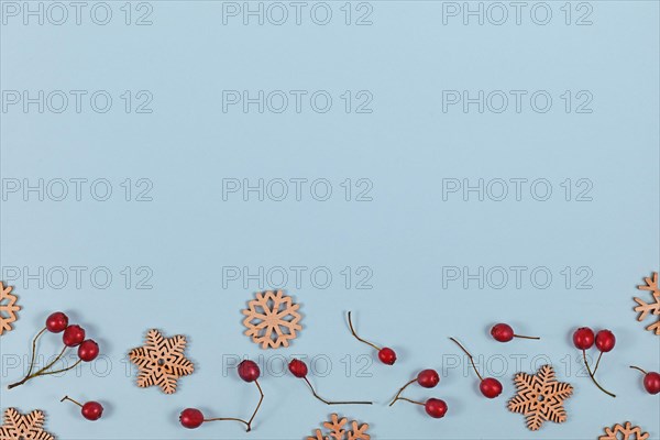 Seasonal flat lay with red winter berries and wooden snowflake Christmas ornaments at bottom of light blue background with empty copy space