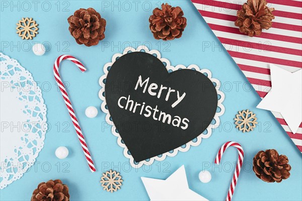 Christmas flat lay with heart shaped chalkboard with text Merry Christmas on light blue background with seasonal decoration like candy cane