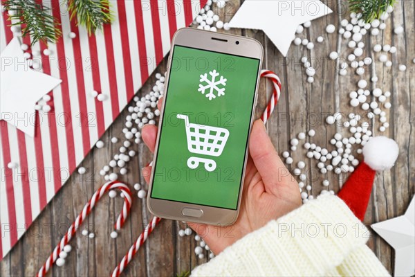 Concept for Christmas seasonal online gift shopping with hand holding cell phone with white shopping cart sign on green background in front of wooden desk with seasonal decoration