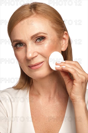 Portrait of a beautiful elderly woman in a white shirt removing makeup with a cleanser and a cotton pad