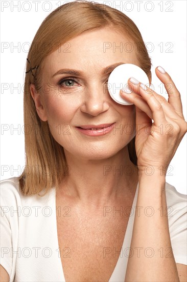 Portrait of a beautiful elderly woman in a white shirt removing makeup with a cleanser and a cotton pad