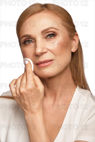 Portrait of a beautiful elderly woman in a white shirt removing makeup with a cleanser and a cotton pad