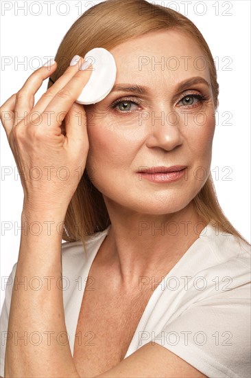 Portrait of a beautiful elderly woman in a white shirt removing makeup with a cleanser and a cotton pad