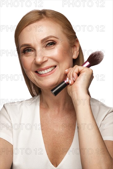 Portrait of a beautiful elderly woman in a white shirt with classic makeup and blond hair with cosmetic brush in her hands