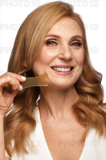 Portrait of a beautiful elderly woman in a white shirt with classic makeup and blond hair with massage tools in hand