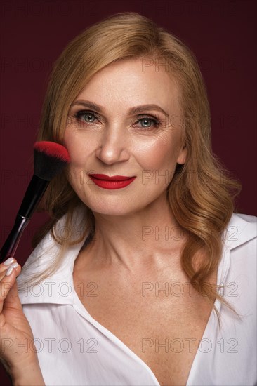 Portrait of a beautiful elderly woman in a white shirt with classic makeup and blond hair with a face brush in hand