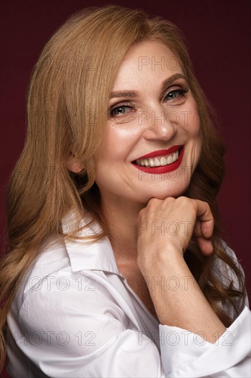 Portrait of a beautiful elderly woman in a white shirt with classic makeup and blond hair with red lipstick in hand