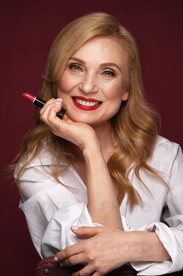 Portrait of a beautiful elderly woman in a white shirt with classic makeup and blond hair with red lipstick in hand