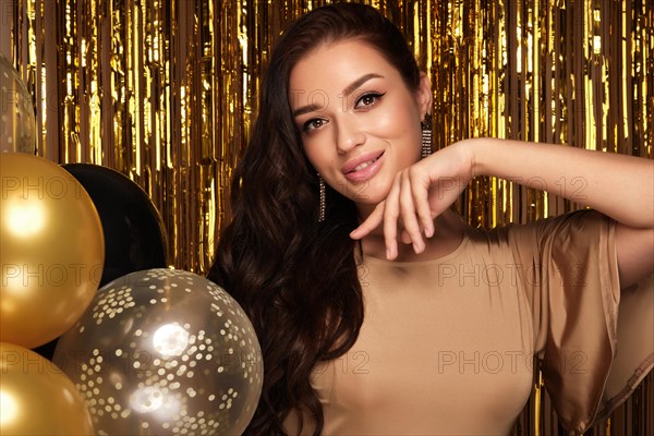 Beautiful young girl in elegant evening dress with festive balloons on the background of gold decor