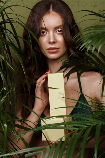 Beautiful sexy woman in a bikini posing in the middle of green plants with a box of cosmetics in hand. Summer look. Beauty face. Photo taken in the studio