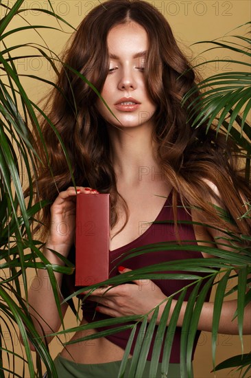 Beautiful sexy woman in a bikini posing in the middle of green plants with a box of cosmetics in hand. Summer look. Beauty face. Photo taken in the studio