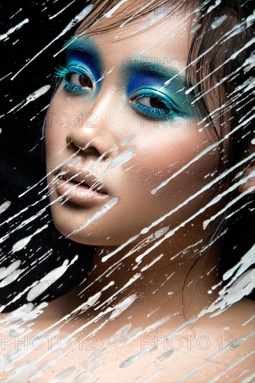 Beautiful Asian girl with bright blue make-up behind glass with drops of wax. Beauty face. Picture taken in the studio on a black background