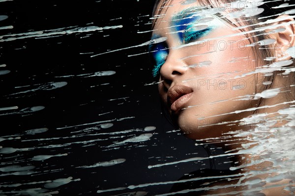 Beautiful Asian girl with bright blue make-up behind glass with drops of wax. Beauty face. Picture taken in the studio on a black background