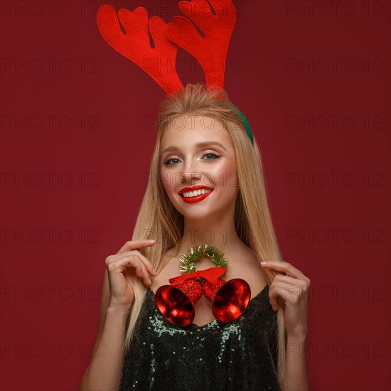 Beautiful blonde girl in a New Year's image with Christmas bells around her neck and deer horns on her head. Beauty face with festive makeup. Photo taken in the studio