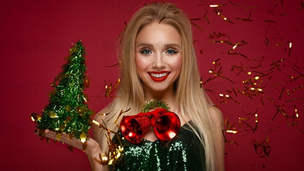 Beautiful blonde girl in a New Year's image with Christmas bells around her neck and green tree. Beauty face with festive makeup. Photo taken in the studio
