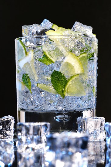 Glass of Mojito summer alcoholic cocktail with ice cubes mint and lime on black background with raw lime and mint leaf