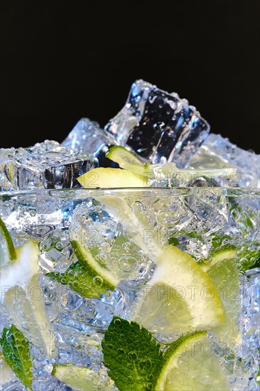 Glass of Mojito summer alcoholic cocktail with ice cubes mint and lime on black background with raw lime and mint leaf